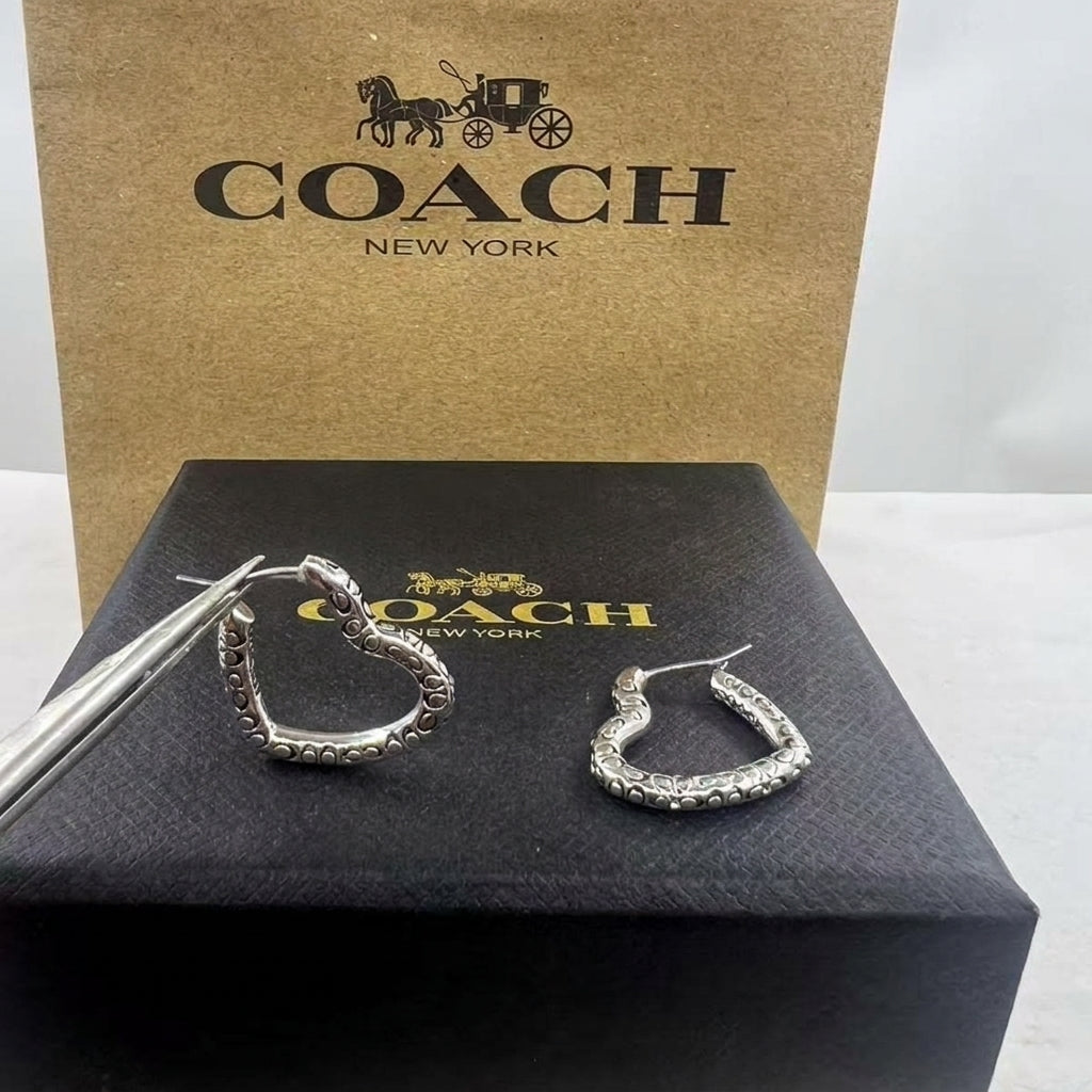 A pair of silver-tone Coach hoop earrings with pavé-set clear stones, shown with metal tweezers on top of a black Coach New York box, with a larger brown Coach New York box behind it.