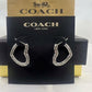 A pair of silver-tone hoop earrings with textured, scalloped edges, displayed on a black Coach New York card in front of a gold and black Coach box.