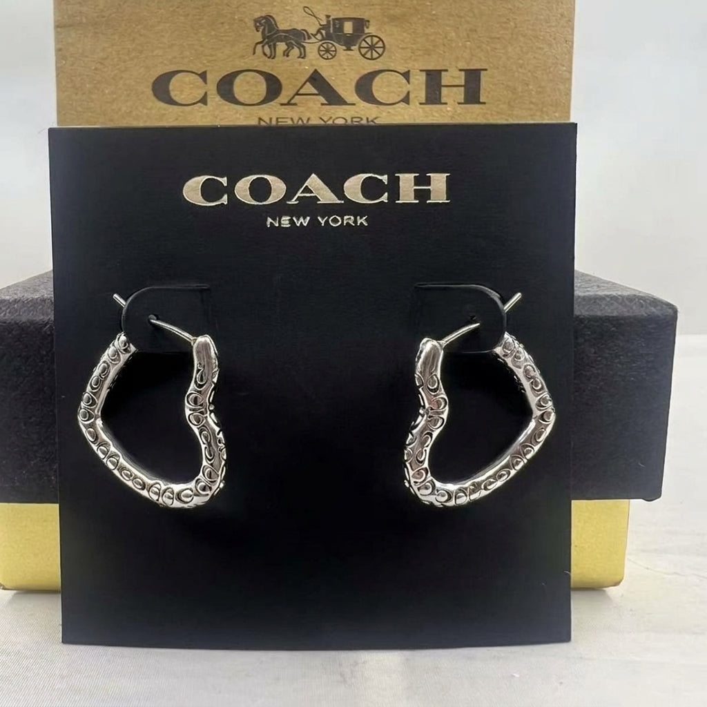 A pair of silver-tone hoop earrings with textured, scalloped edges, displayed on a black Coach New York card in front of a gold and black Coach box.