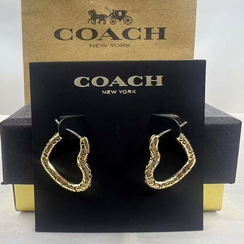 A pair of gold-tone Coach huggie hoop earrings displayed on a black Coach New York card, with the brown Coach box behind it on a white background.