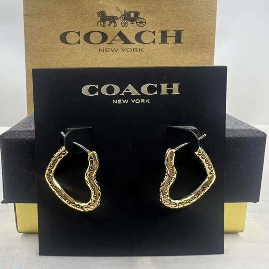 A pair of gold-tone Coach huggie hoop earrings displayed on a black Coach New York card, with the brown Coach box behind it on a white background.