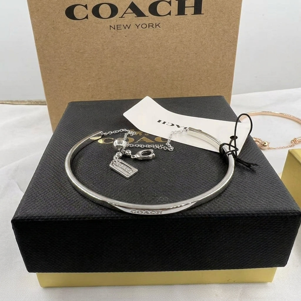 A silver-tone Coach bangle bracelet with a signature logo charm, displayed on a black textured Coach box with its signature kraft paper box and hangtag behind it.