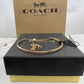 A gold-tone Coach charm bracelet displayed on a black textured box with a gold-tone base, in front of a brown Coach New York box.
