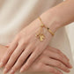 A delicate gold bracelet with dangling charms, including a small heart, a tag, and a tiny cross, worn on a woman's wrist. The bracelet also features a smooth gold band.