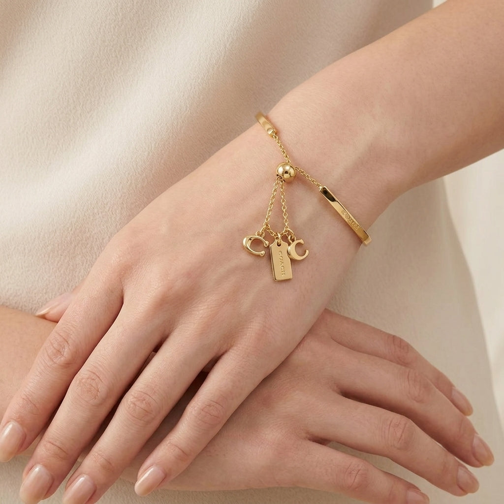 A delicate gold bracelet with dangling charms, including a small heart, a tag, and a tiny cross, worn on a woman's wrist. The bracelet also features a smooth gold band.
