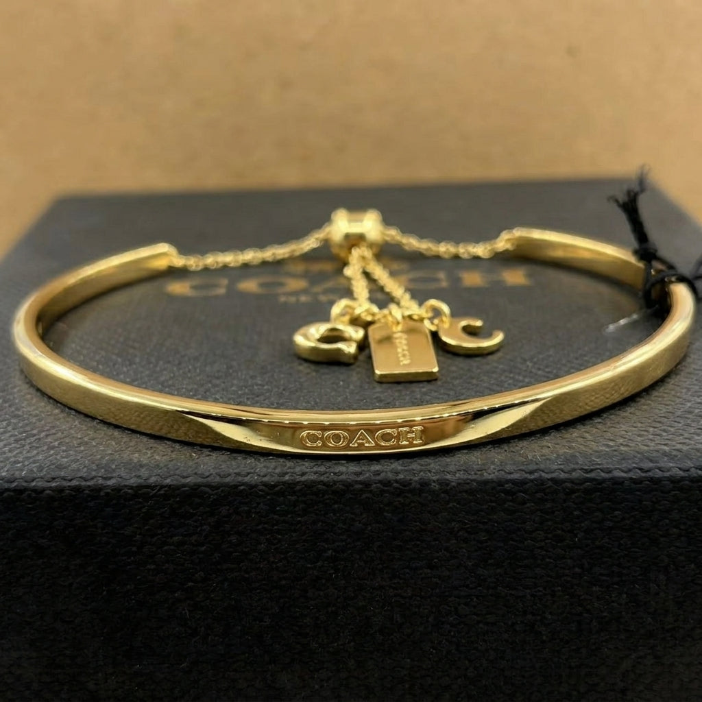 A gold-tone Coach bangle bracelet with a chain extender and three charms (Coach logo, 'COACH' tag, and 'C' charms), resting on a black textured box with the Coach logo visible.