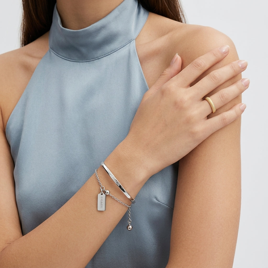 A close-up of a woman wearing a silver bangle bracelet and a matching silver charm bracelet, paired with a gold band on her finger, while wearing a light blue satin halter top.