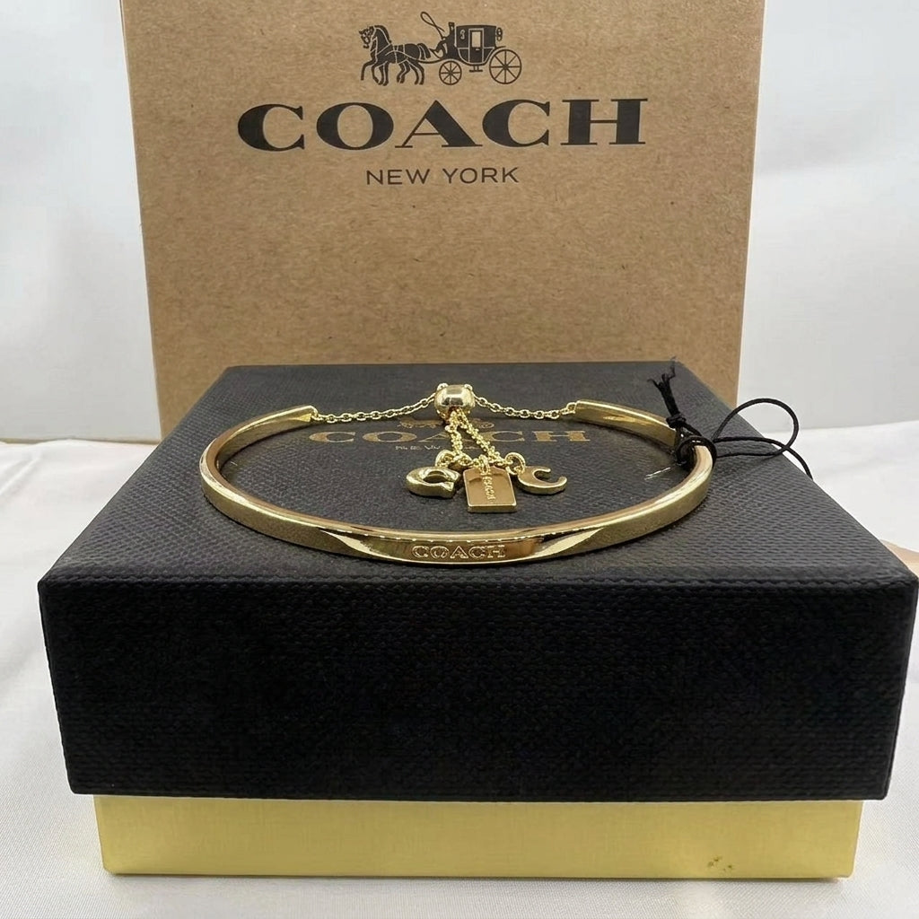 A gold-tone Coach bangle bracelet with a padlock charm and horse-carriage pendant, displayed on a black Coach presentation box with a brown Coach box behind it.