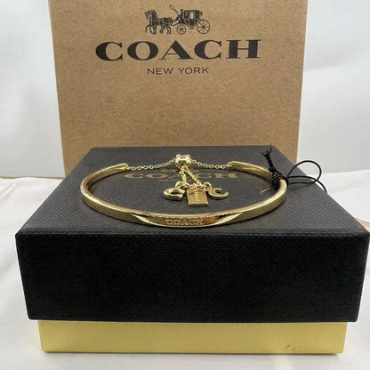 A gold-tone Coach bangle bracelet with a padlock charm and horse-carriage pendant, displayed on a black Coach presentation box with a brown Coach box behind it.
