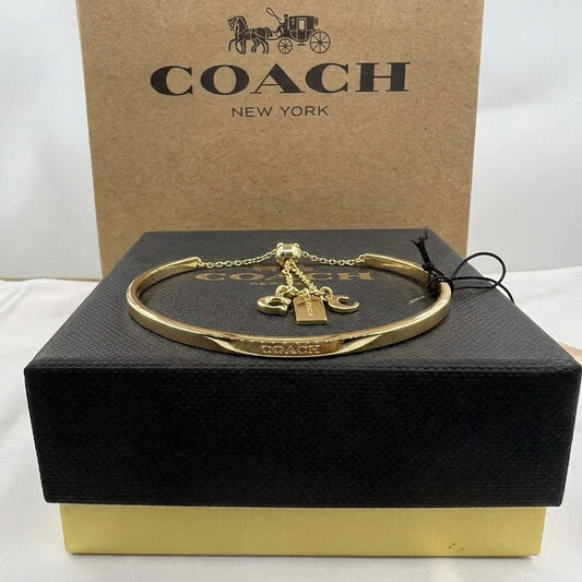 A gold-tone Coach bangle bracelet with a padlock charm and horse-carriage pendant, displayed on a black Coach presentation box with a brown Coach box behind it.