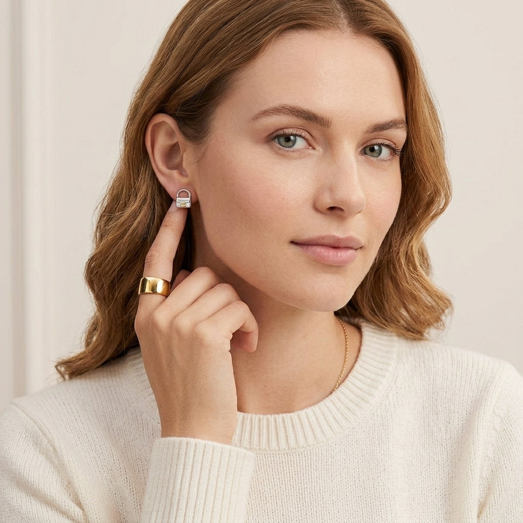 A close-up of a woman wearing a small silver and white geometric stud earring, with a gold ring on one finger and a delicate gold necklace. She is wearing a cream-colored ribbed knit sweater against a soft, light beige background.