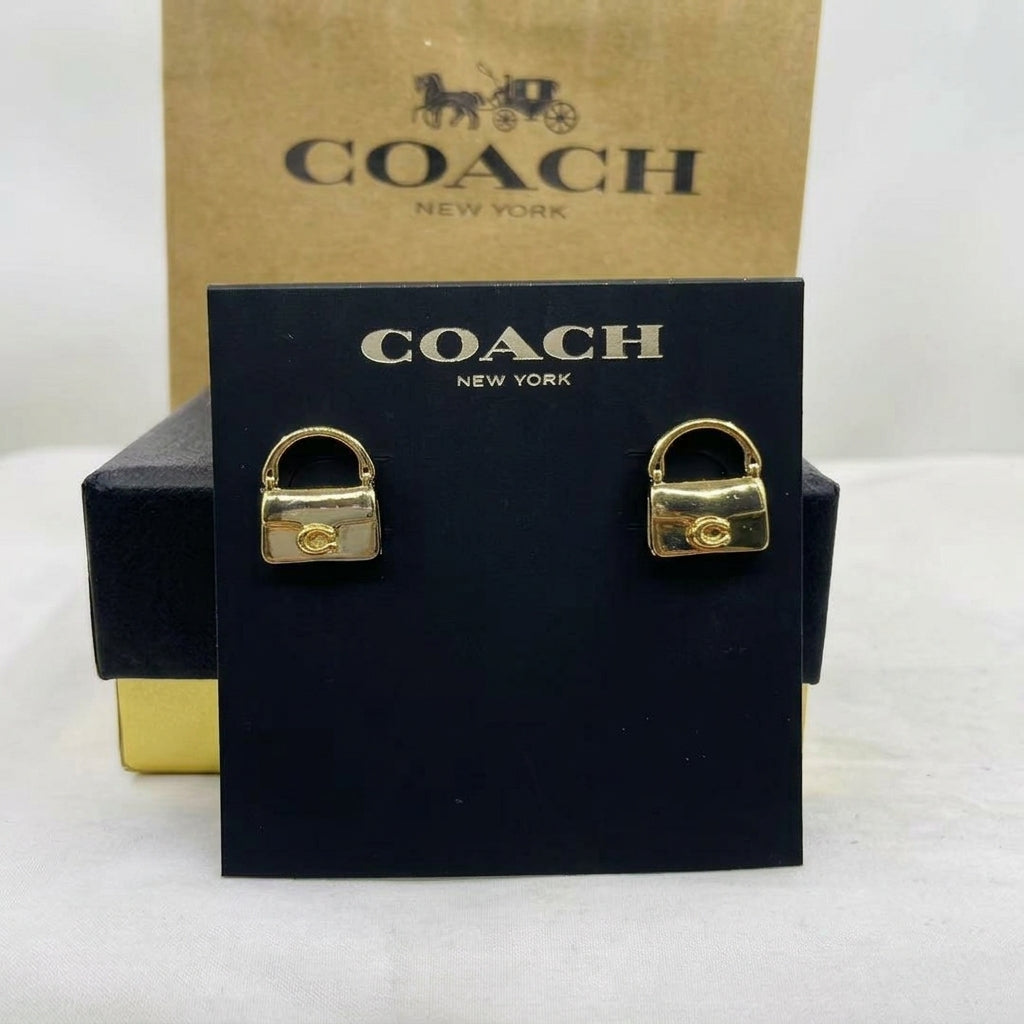 A pair of gold-tone Coach New York stud earrings shaped like small padlocks, displayed on a black Coach New York card with the brand's brown box behind it.