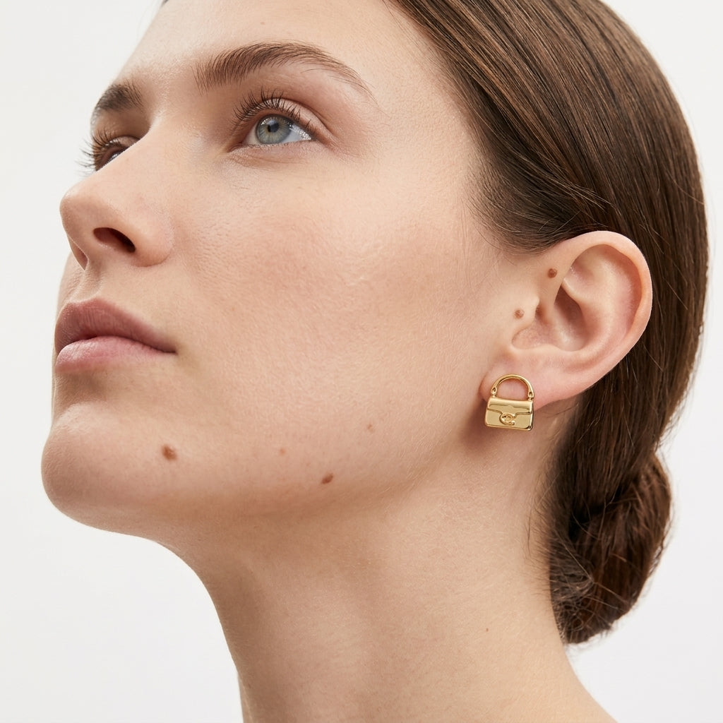 A close-up of a woman wearing a small gold stud earring shaped like a padlock with a tiny keyhole detail, shown against a clean light gray background.