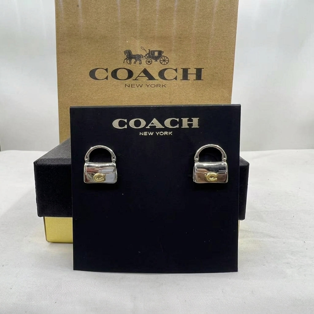 A pair of Coach padlock stud earrings displayed on a black branded card, with a brown Coach box behind them on a white surface.