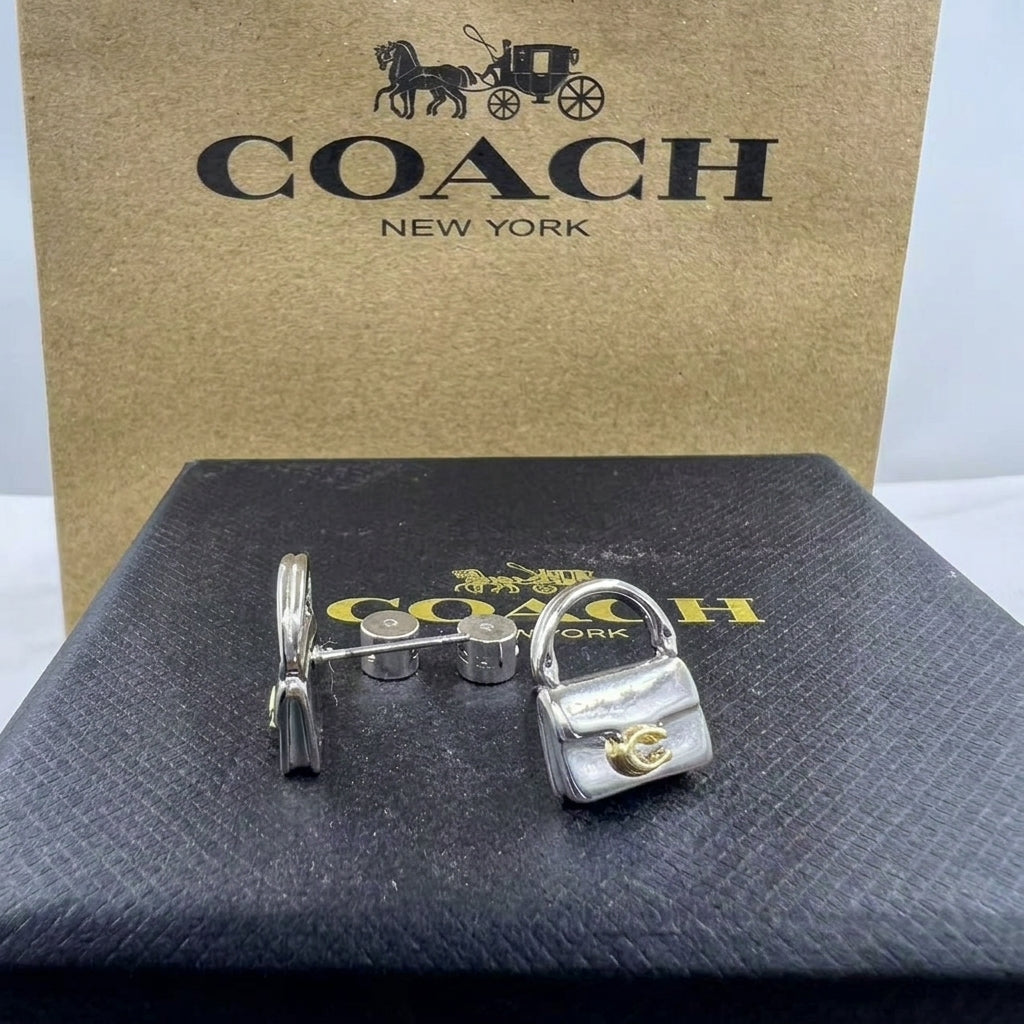 A pair of Coach New York cufflinks shaped like a small silver handbag and a silver padlock, displayed on a black textured Coach box with the gold Coach New York logo, in front of a brown Coach box.