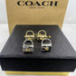 A pair of gold and silver padlock-shaped earrings displayed on a black textured Coach box, with a brown Coach New York box in the background.