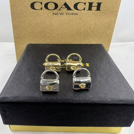 A pair of gold and silver padlock-shaped earrings displayed on a black textured Coach box, with a brown Coach New York box in the background.