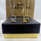 Two Coach lock-shaped pendant necklaces, one silver and one gold, displayed on top of a black and gold presentation box with a Coach New York box in the background.