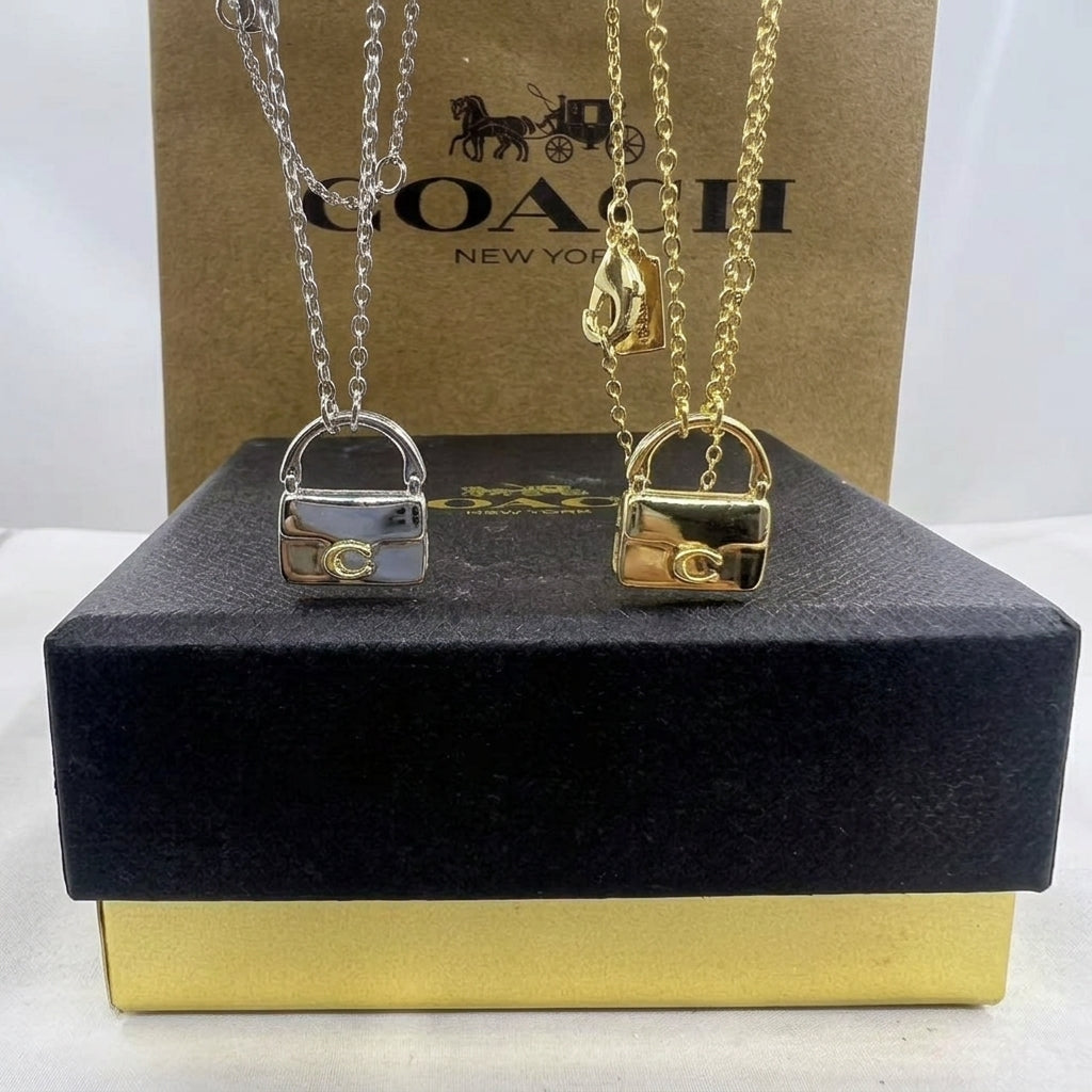 Two Coach lock-shaped pendant necklaces, one silver and one gold, displayed on top of a black and gold presentation box with a Coach New York box in the background.