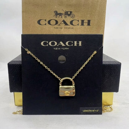 A gold-tone Coach New York padlock necklace displayed on a black branded card, with a brown Coach box behind it on a light neutral background.