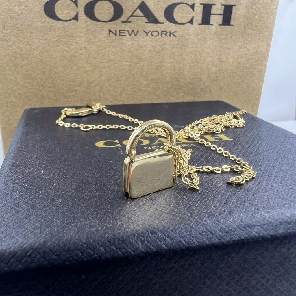 A gold-tone padlock pendant necklace on a delicate chain, displayed on a dark blue Coach box with a brown Coach box in the background.