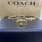 A gold-tone Coach mini handbag charm pendant necklace displayed on a dark blue textured box, with the signature Coach New York box in the background.