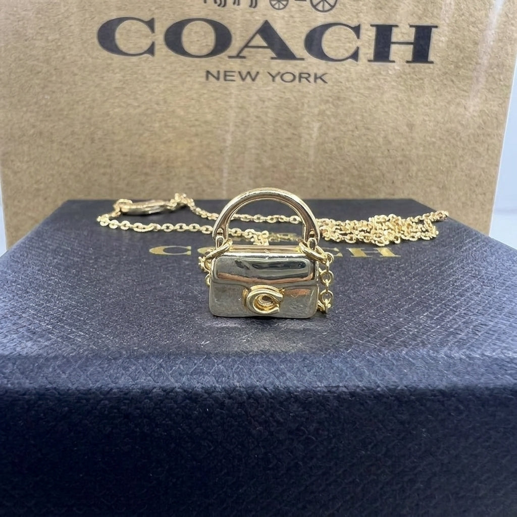 A gold-tone Coach mini handbag charm pendant necklace displayed on a dark blue textured box, with the signature Coach New York box in the background.