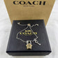 A Coach New York silver-tone key and padlock charm bracelet displayed on its black presentation box, with the signature gold-tone Coach box behind it.