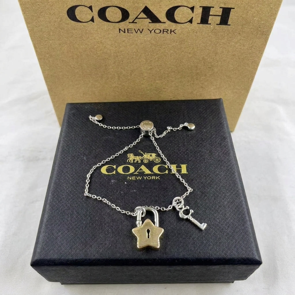 A Coach New York silver-tone key and padlock charm bracelet displayed on its black presentation box, with the signature gold-tone Coach box behind it.