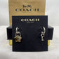A pair of Coach New York earrings displayed on a black branded card, with a gold-tone star-shaped earring and a silver-tone key-shaped earring, in front of a Coach box.