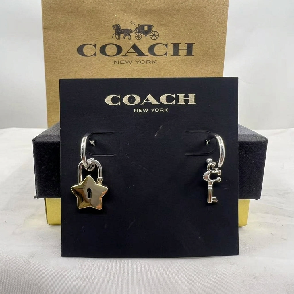 A pair of Coach New York earrings displayed on a black branded card, with a gold-tone star-shaped earring and a silver-tone key-shaped earring, in front of a Coach box.