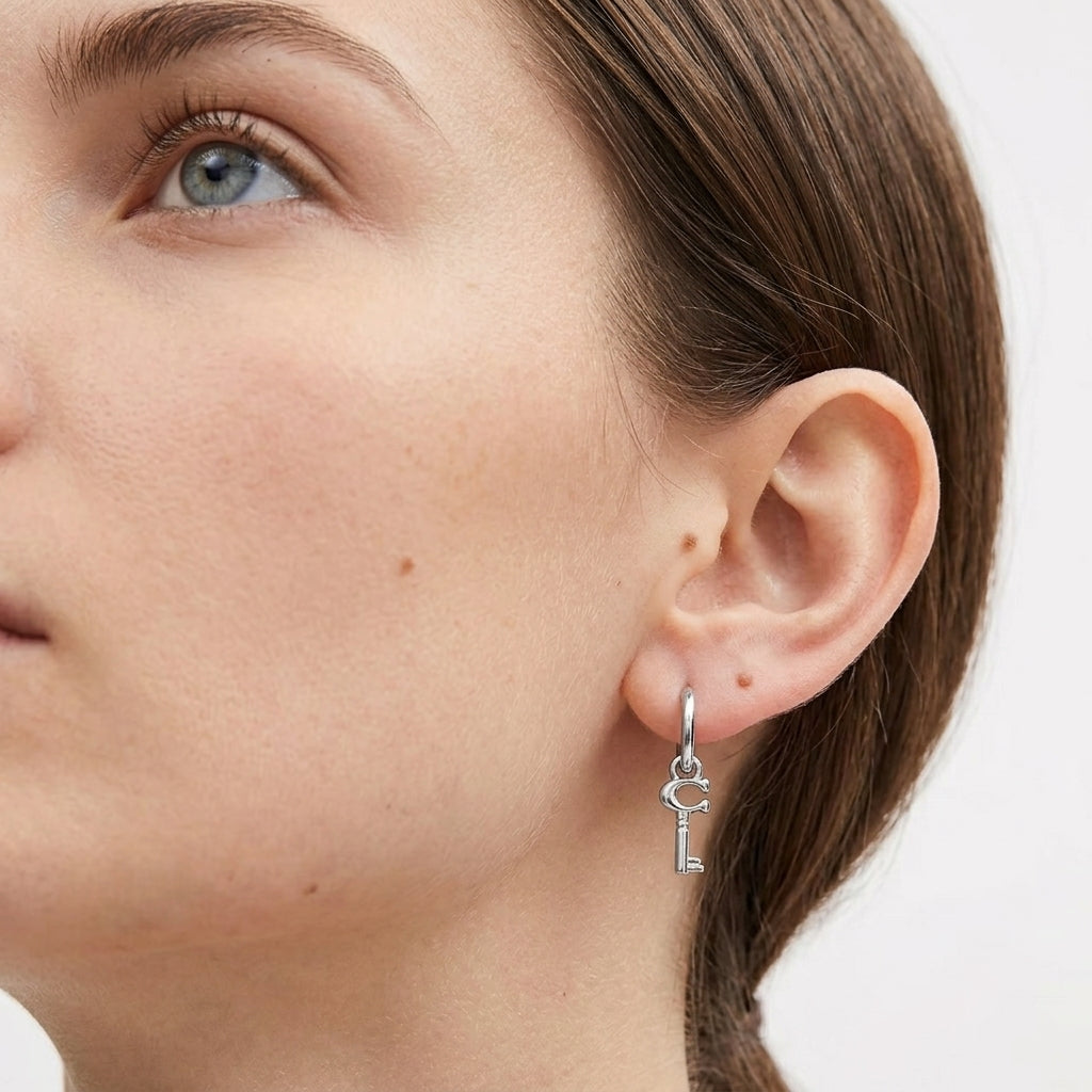 A close-up of a woman's ear wearing a silver key-shaped hoop earring against a clean white background.
