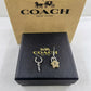 A pair of Coach New York dangle earrings displayed on a black branded box, with a tan Coach New York box behind them. The earrings feature a silver-tone key charm and a gold-tone star-shaped lock charm.