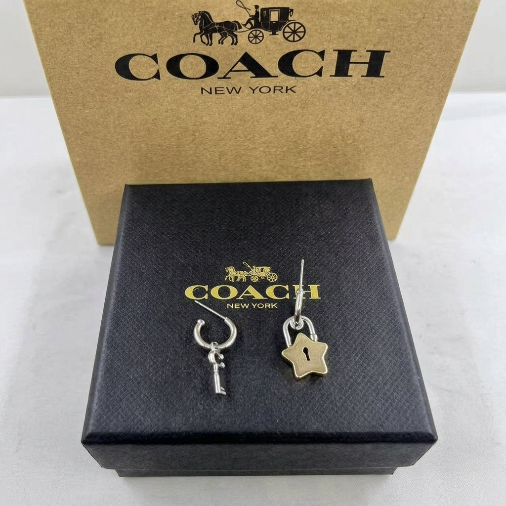 A pair of Coach New York dangle earrings displayed on a black branded box, with a tan Coach New York box behind them. The earrings feature a silver-tone key charm and a gold-tone star-shaped lock charm.