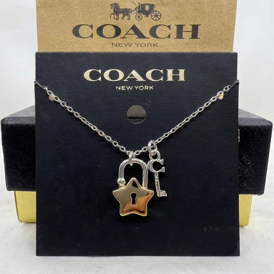A Coach New York necklace with a silver chain, featuring a gold star-shaped padlock and a silver key charm, displayed on its black and brown branded packaging.