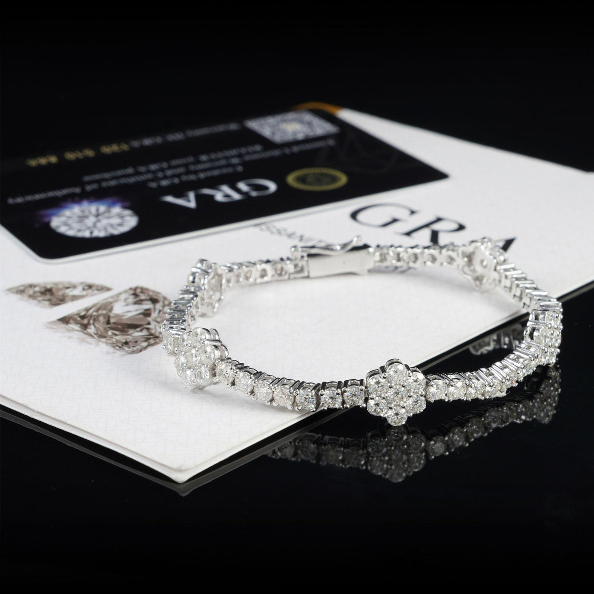 A silver-toned diamond tennis bracelet with a floral cluster centerpiece, displayed on top of a GRA certification card and paper on a glossy black surface.