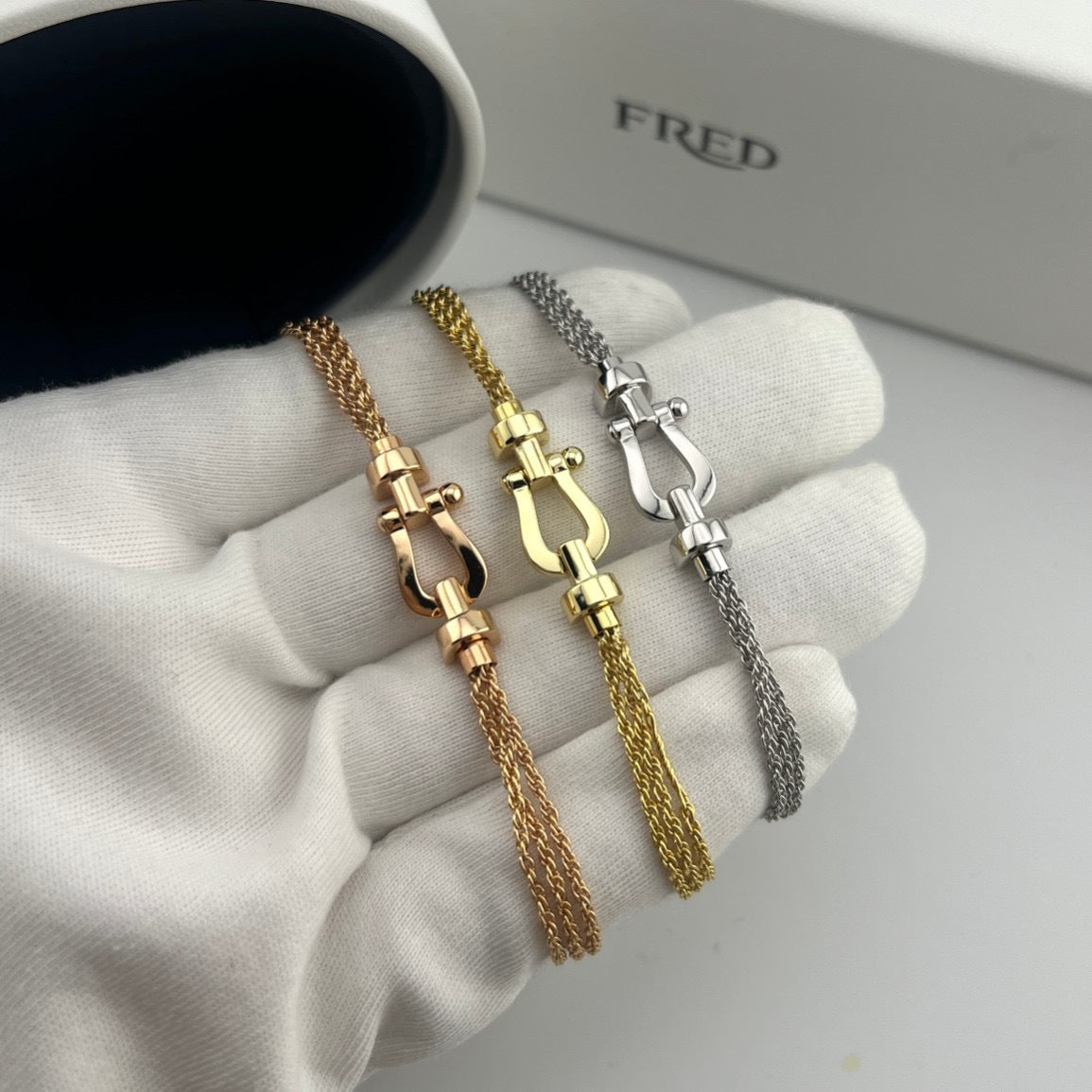 Three gold chain bracelets on a white hand with 'FRED' branding in the background.