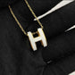 High-End “H” Enamel Pendant Necklace Gold necklace with a white 'H' pendant on a black fabric background