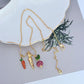 Necklace on a rustic wood or green linen background, highlighting the colorful veggie charm. Alt: “Kate Spade Harvest Time Veggie Charm Pendant Necklace – Playful Garden Jewelry.”