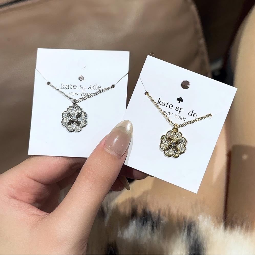 Clean white or soft pastel background — the Kate Spade Heritage Bloom mini pendant displayed on a bust form. The floral enamel pendant catches soft light, showing delicate petal details and the signature Kate Spade aesthetic.