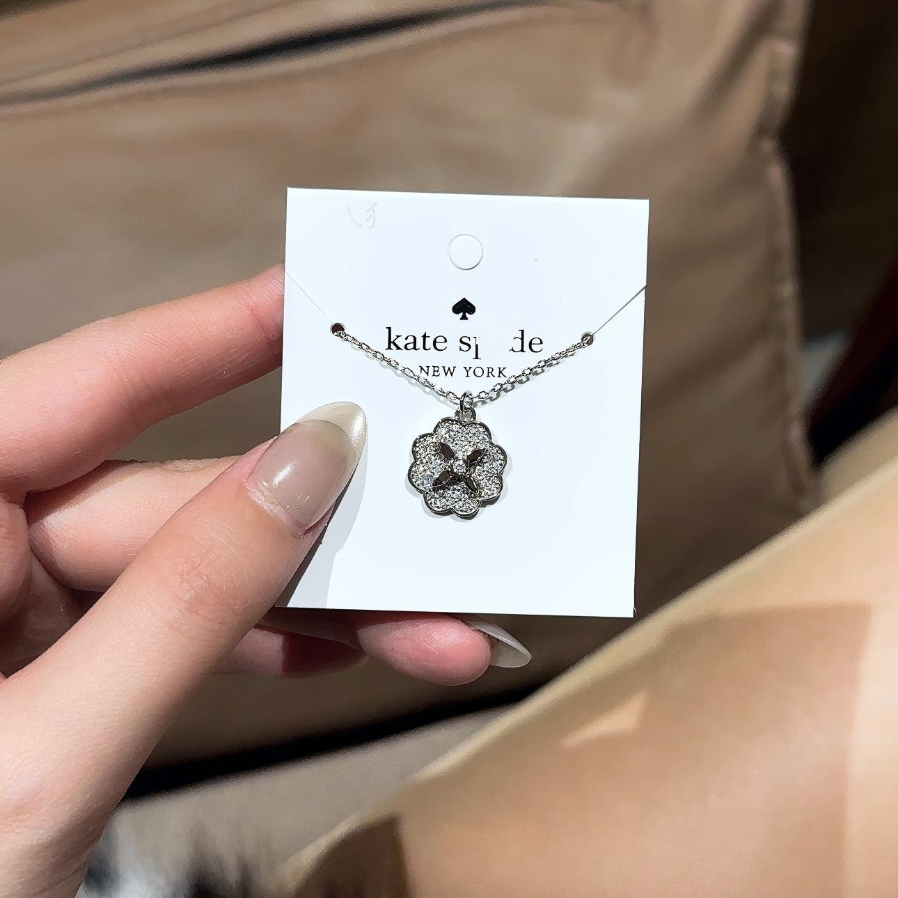 Clean white or soft pastel background — the Kate Spade Heritage Bloom mini pendant displayed on a bust form. The floral enamel pendant catches soft light, showing delicate petal details and the signature Kate Spade aesthetic.