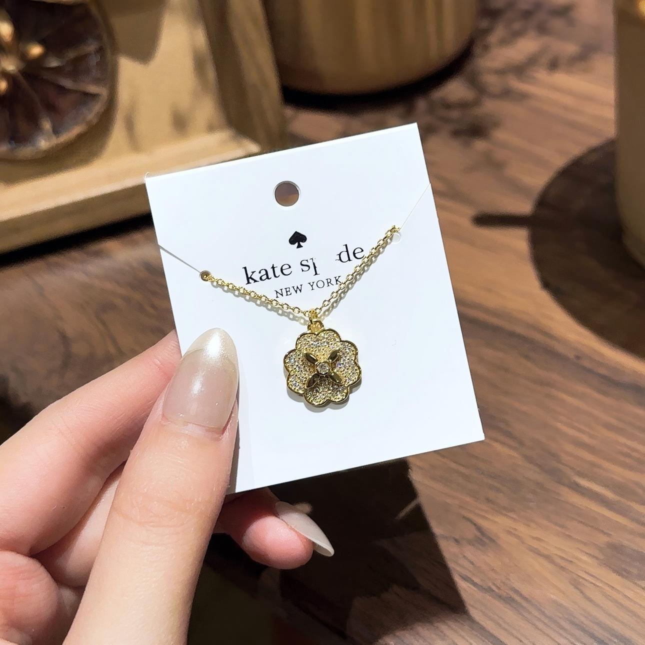 Clean white or soft pastel background — the Kate Spade Heritage Bloom mini pendant displayed on a bust form. The floral enamel pendant catches soft light, showing delicate petal details and the signature Kate Spade aesthetic.