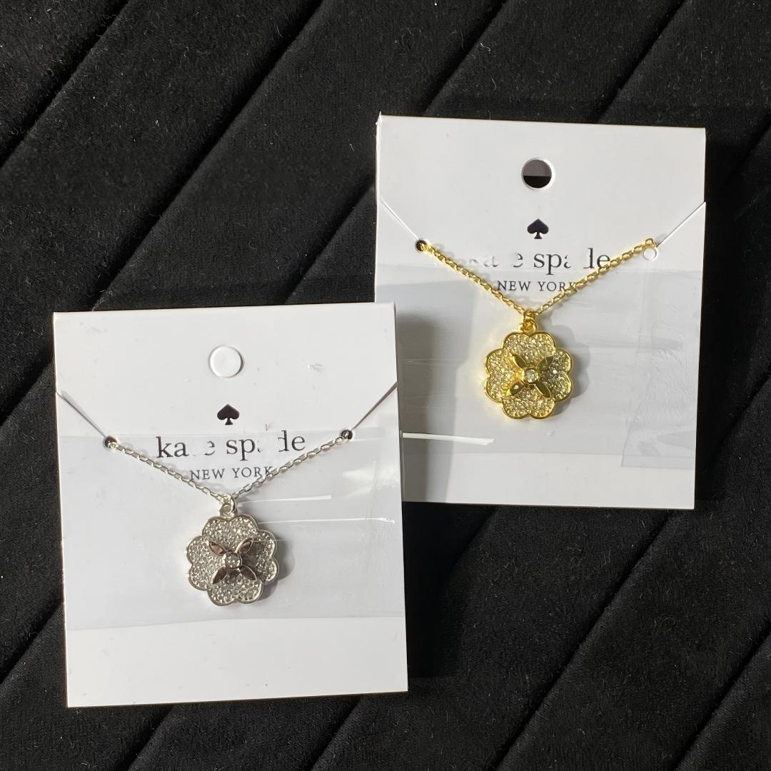 Clean white or soft pastel background — the Kate Spade Heritage Bloom mini pendant displayed on a bust form. The floral enamel pendant catches soft light, showing delicate petal details and the signature Kate Spade aesthetic.