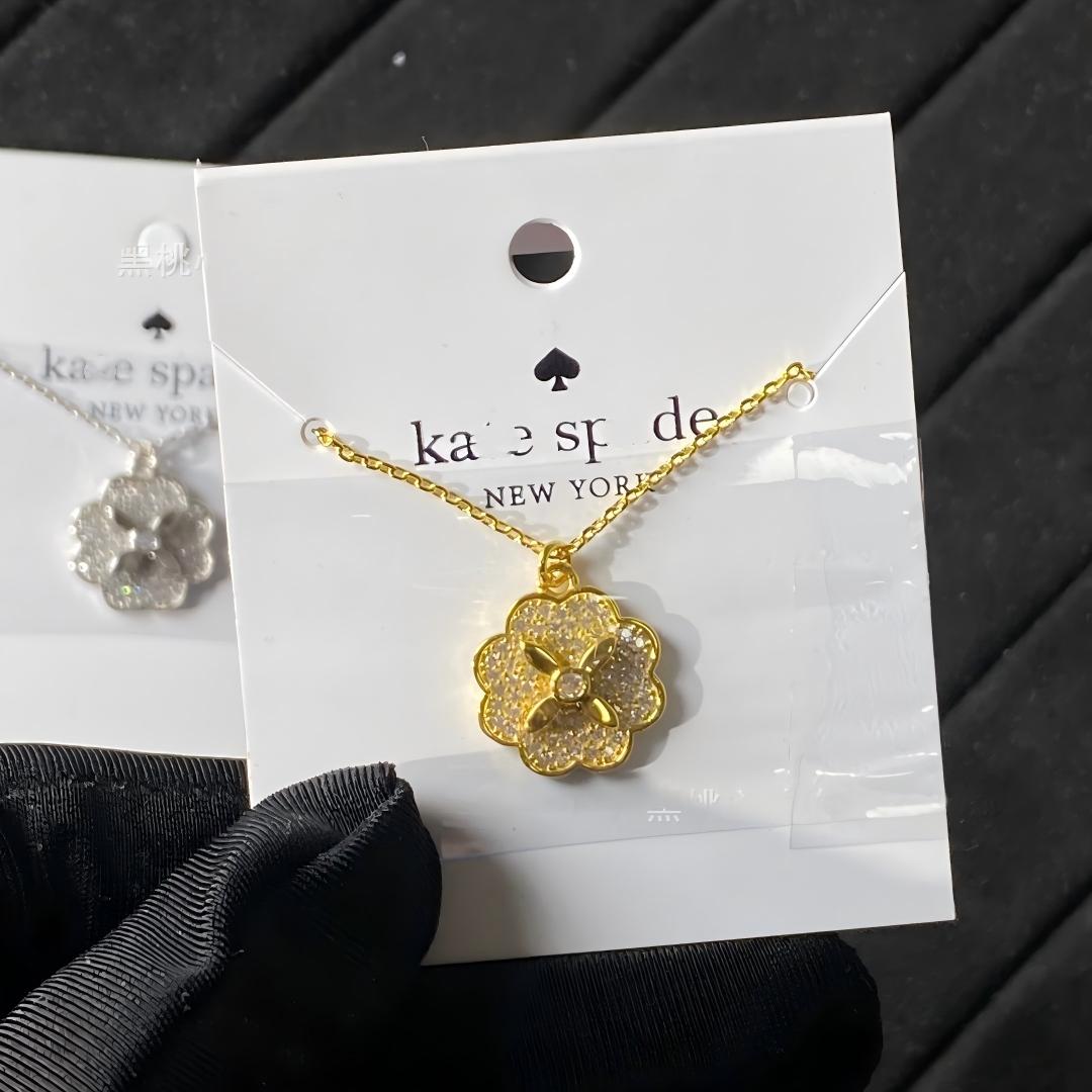 Clean white or soft pastel background — the Kate Spade Heritage Bloom mini pendant displayed on a bust form. The floral enamel pendant catches soft light, showing delicate petal details and the signature Kate Spade aesthetic.