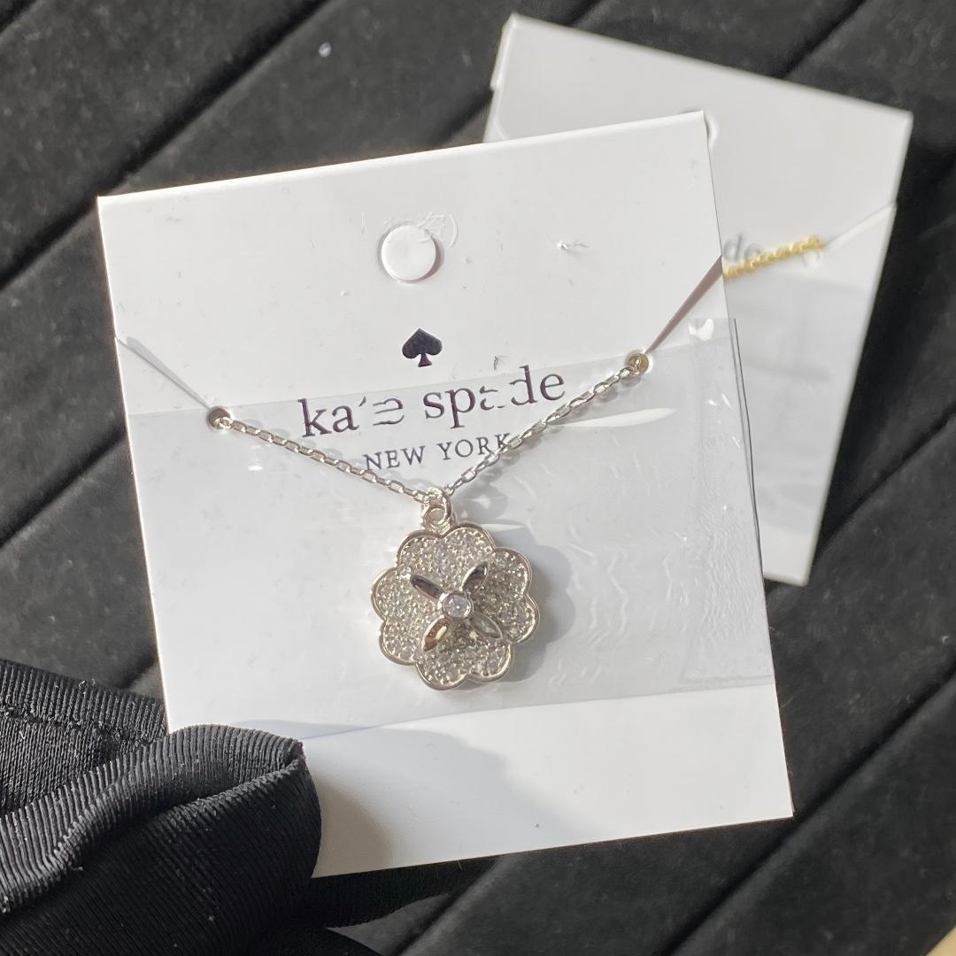 Clean white or soft pastel background — the Kate Spade Heritage Bloom mini pendant displayed on a bust form. The floral enamel pendant catches soft light, showing delicate petal details and the signature Kate Spade aesthetic.