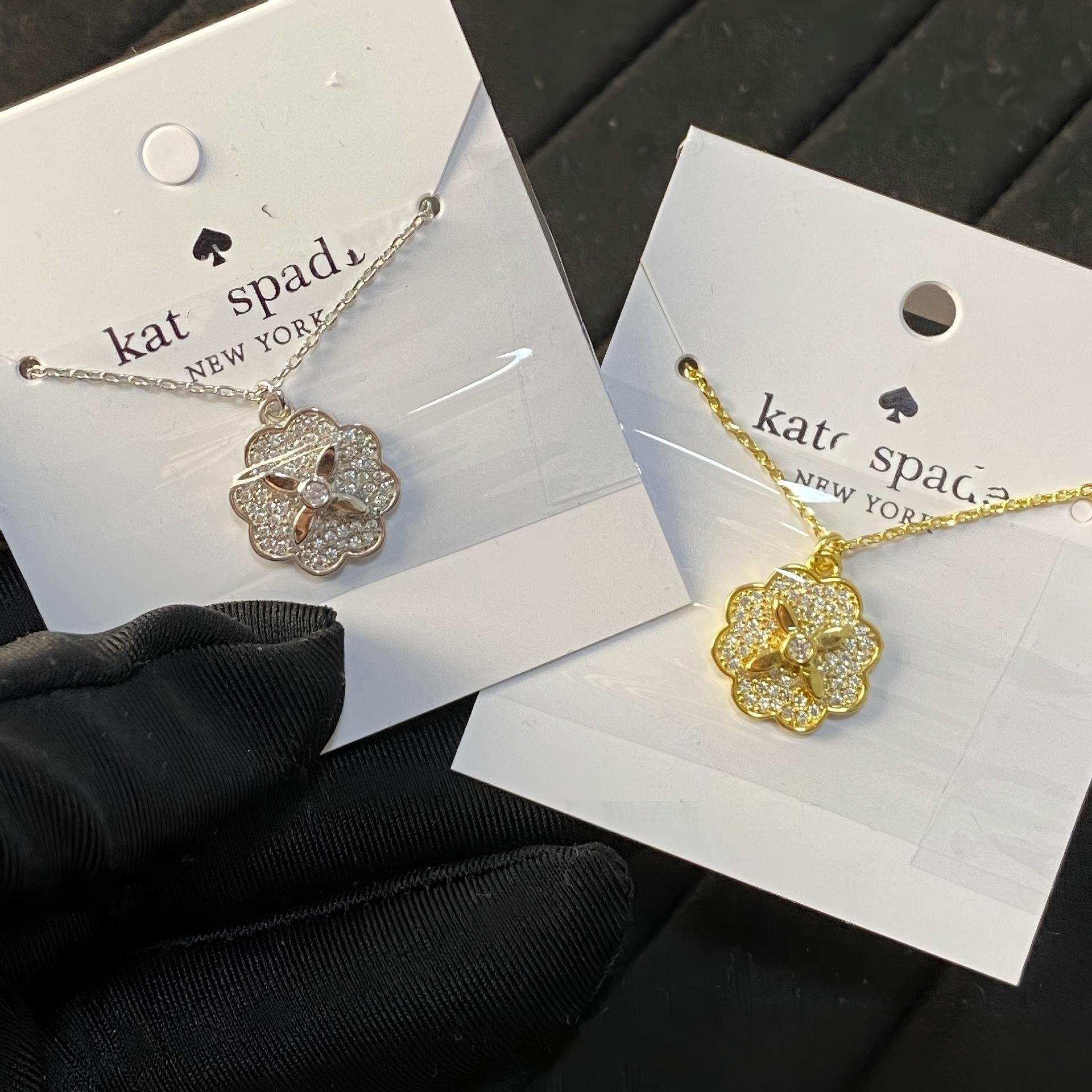 Clean white or soft pastel background — the Kate Spade Heritage Bloom mini pendant displayed on a bust form. The floral enamel pendant catches soft light, showing delicate petal details and the signature Kate Spade aesthetic.