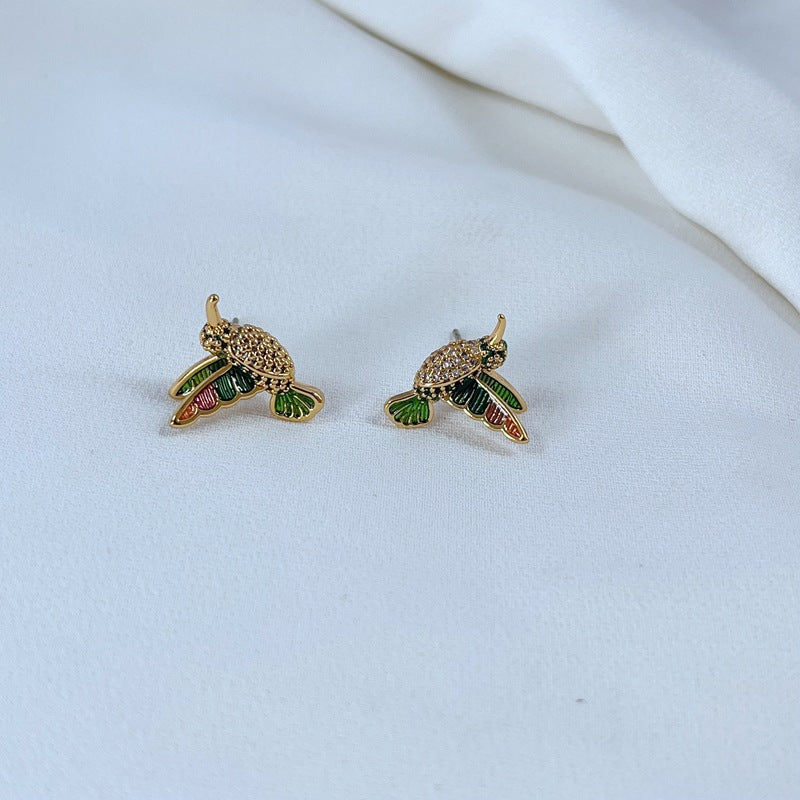 Earrings on a soft sage green or misty blue background, showcasing the hummingbird charm with iridescent enamel wings. Alt: “Kate Spade New York Hummingbird Earrings – Enamel Bird Drop Jewelry.”