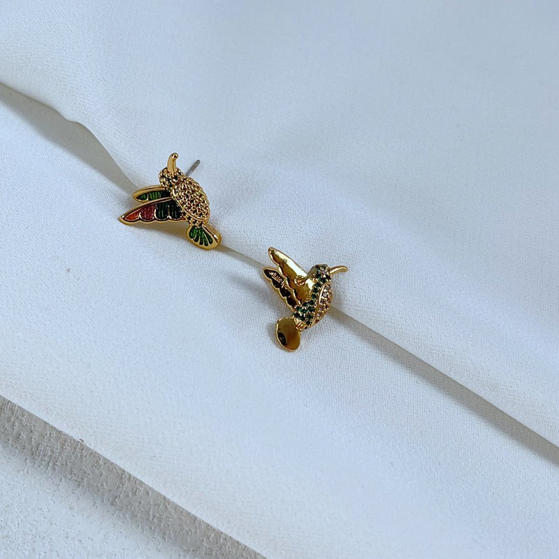 Earrings on a soft sage green or misty blue background, showcasing the hummingbird charm with iridescent enamel wings. Alt: “Kate Spade New York Hummingbird Earrings – Enamel Bird Drop Jewelry.”