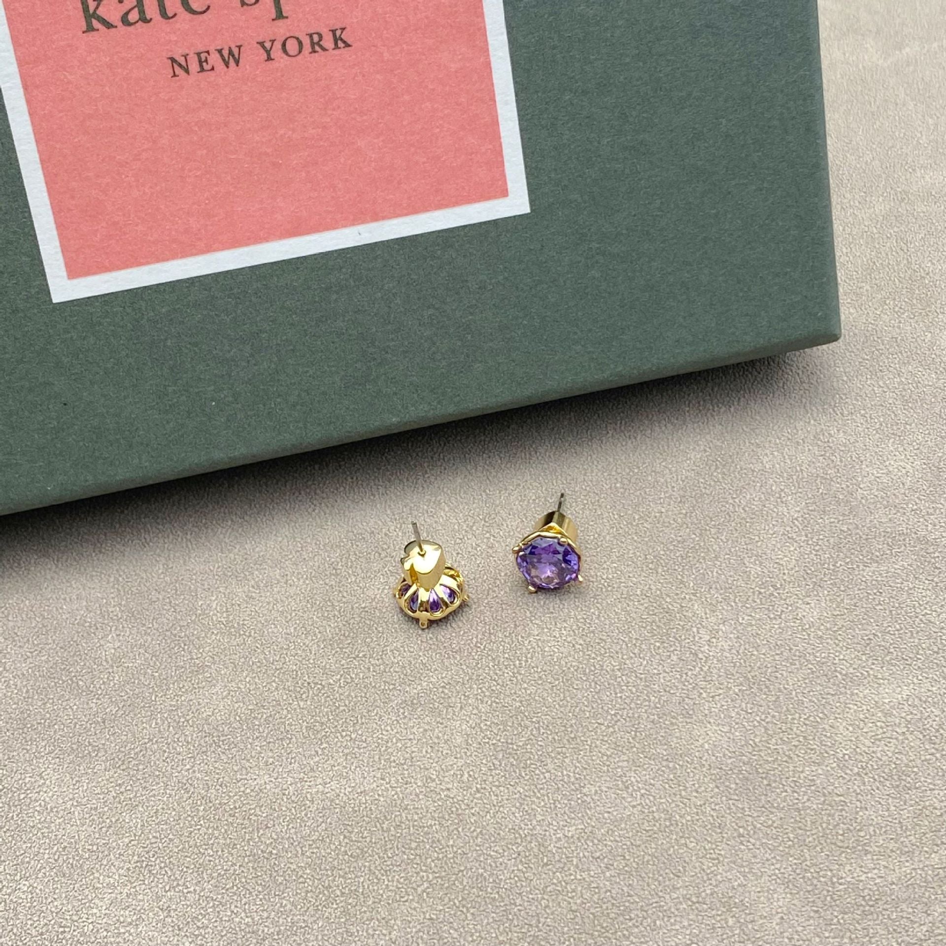 Gold and purple earrings on a gray surface with a Kate Spade box in the background.