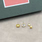 Gold and silver stud earrings on a gray surface with a pink and green box in the background.